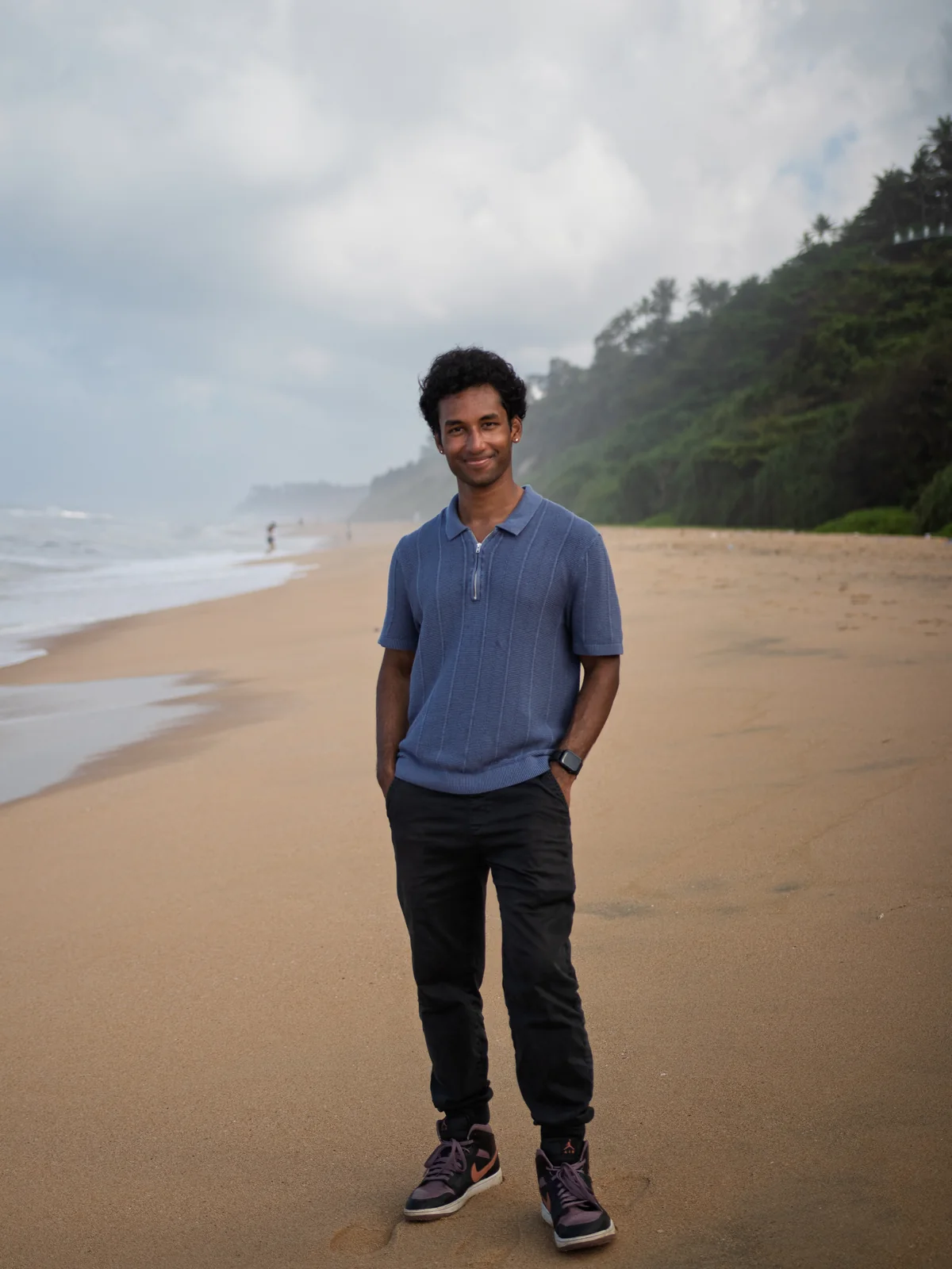Sai standing on the beach at Varkala in a blue polo, smiling, cliffs and sea behind him